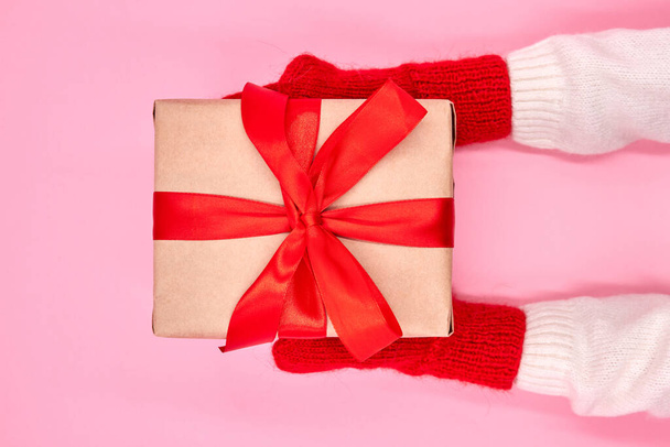 A kesztyűs gyermek karácsonyi vagy újévi ajándékdobozt tart rózsaszín háttér mellett. - Fotó, kép