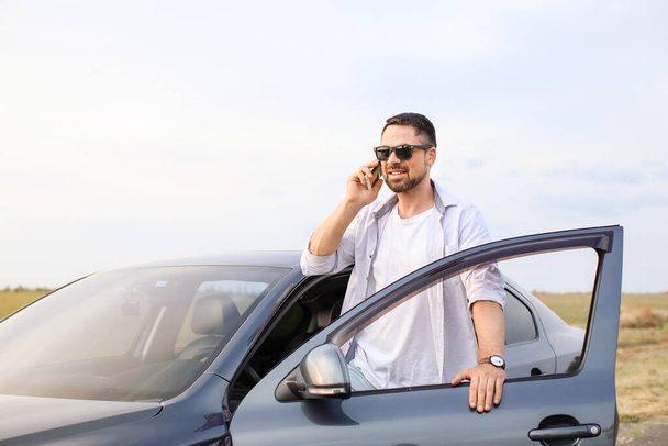 Όμορφος άντρας μιλάει από κινητό κοντά στο μοντέρνο του αυτοκίνητο. - Φωτογραφία, εικόνα