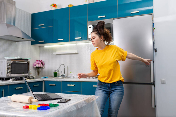 若い美しい女性が冷蔵庫を開き、彼女のラップトップを注意深く見ています。屋内だ。オンラインのレシピやビデオ通信を使って自宅で調理するというコンセプト. - 写真・画像
