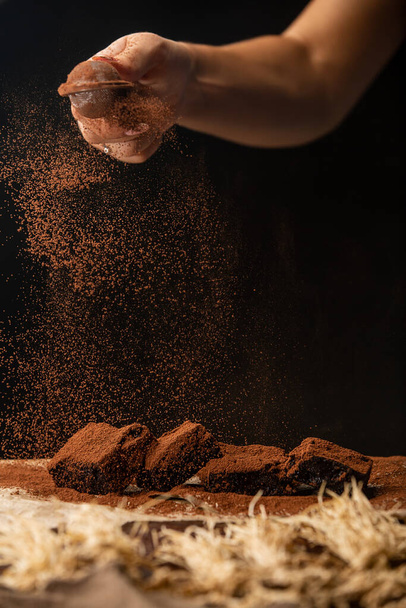 Detailní záběr na ruku je sypání kakaa prášek na čokoládové brownies na pečení papíru na černém pozadí - Fotografie, Obrázek
