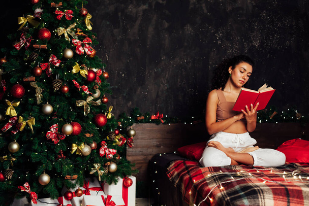 Hermosa mujer lee un libro en la cama en el árbol de Navidad con regalos de año nuevo - Foto, Imagen