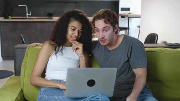 Sorprendida pareja mixta leyendo grandes noticias en el ordenador portátil, el hombre joven y la mujer son gratamente sorprendidos mostrando WOW emoción mientras se relaja en el sofá en casa. Emocionado.. - Metraje, vídeo