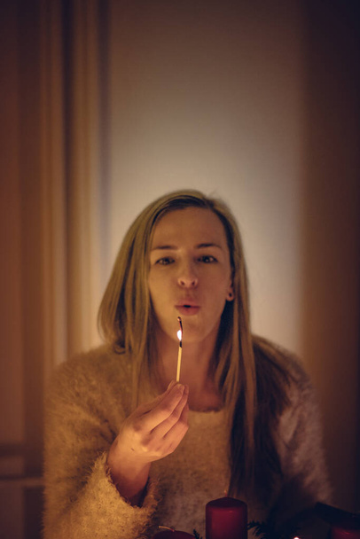 blonde woman in a warm white sweater blows out a candle with which she lit a candlestick. The first Sunday of Advent. Young girl playing with matches. Family time. - Фото, изображение