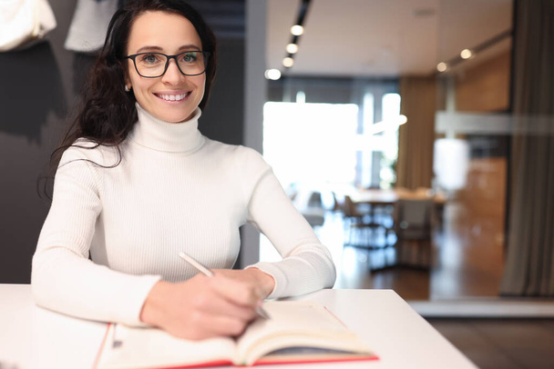 Bruna donna con gli occhiali sorridente e tenendo penna a sfera su notebook - Foto, immagini