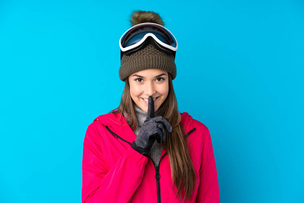 Teenager skier girl with snowboarding glasses over isolated blue background doing silence gesture - Photo, Image