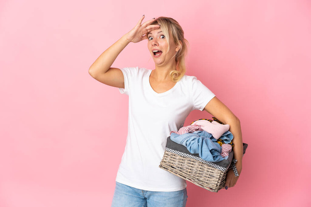 Young Russian woman holding a clothes basket isolated on pink background doing surprise gesture while looking to the side - 写真・画像