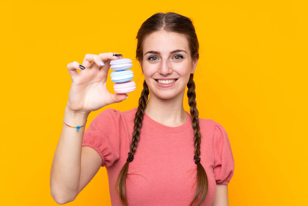 Young woman over isolated yellow background holding colorful French macarons and with happy expression - Photo, Image