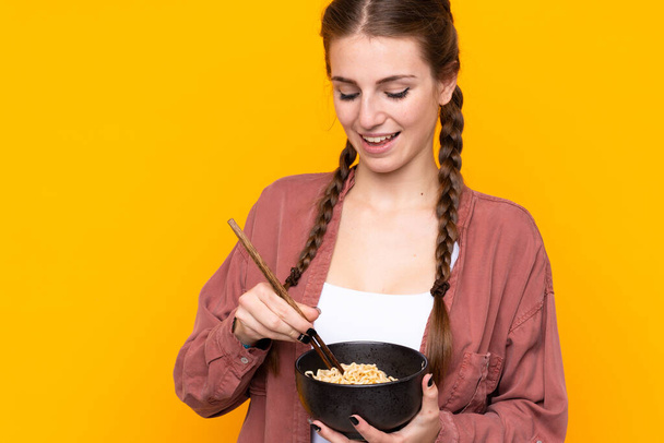 İzole edilmiş sarı arka planda genç bir kadın elinde yemek çubuklarıyla bir kase erişte tutuyor. - Fotoğraf, Görsel