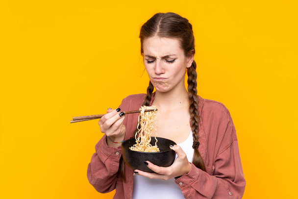Young woman over isolated yellow background holding a bowl of noodles with chopsticks - Valokuva, kuva