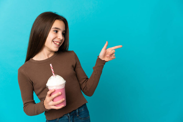 Маленькая девочка с клубничным молочным коктейлем изолирована на синем фоне, указывая пальцем в сторону и представляя продукт - Фото, изображение