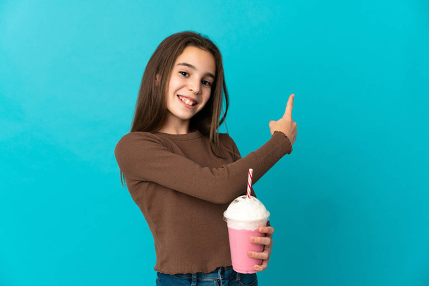 Маленькая девочка с клубничным молочным коктейлем изолирована на синем фоне, указывая назад - Фото, изображение