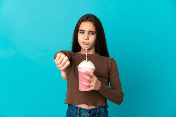 Маленькая девочка с клубничным молочным коктейлем на голубом фоне показывает большой палец вниз с отрицательным выражением - Фото, изображение