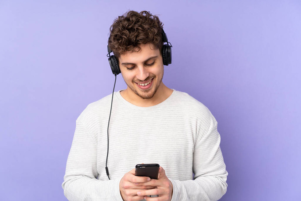 Caucasian man isolated on purple background listening music and looking to mobile - Fotografie, Obrázek