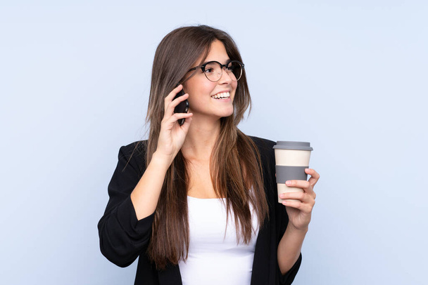 Fiatal brazil üzletasszony kezében kávé, hogy vigye el, és beszél a mobil izolált kék háttér - Fotó, kép
