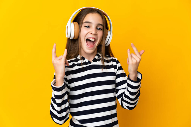 Little girl isolated on yellow background listening music making rock gesture - Fotoğraf, Görsel