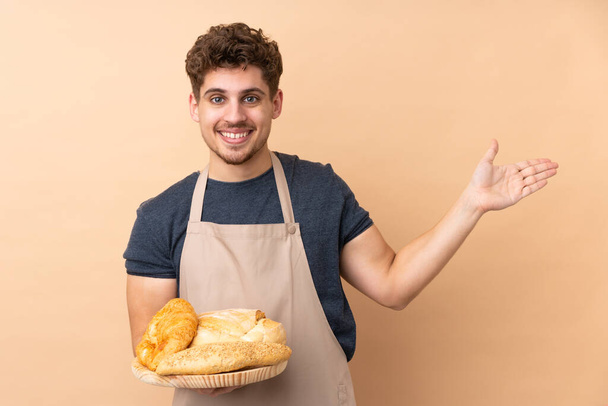 Мужчина пекарь держа стол с несколькими хлебами изолированы на бежевом фоне протягивая руки в сторону для приглашения прийти - Фото, изображение