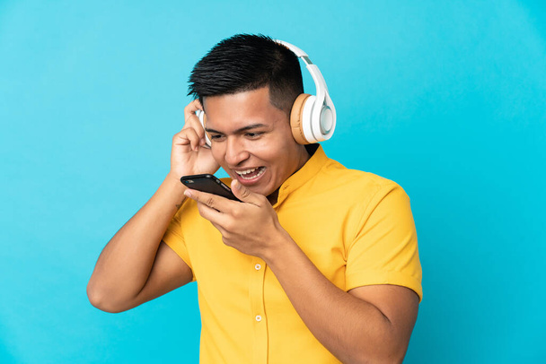 Joven ecuatoriano aislado sobre fondo azul escuchando música con móvil y cantando - Foto, Imagen