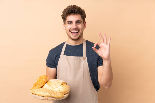 Мужчина пекарь держа стол с несколькими хлебами изолированы на бежевом фоне показывая знак ОК с пальцами - Фото, изображение