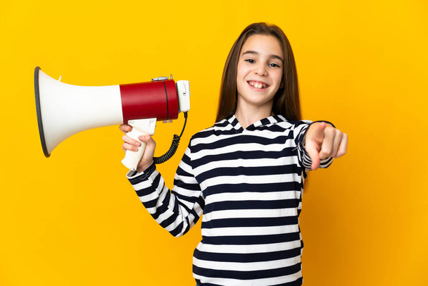 Little girl isolated on yellow background holding a megaphone and smiling while pointing to the front - Valokuva, kuva