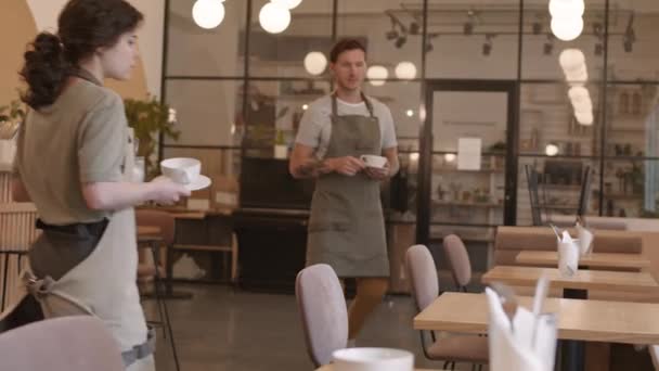 Férfi és női kaukázusi pincérek asztalokat terítenek a vendégek nélküli étteremben nyitás előtt, csészéket tesznek le csészealjakra, különböző irányokba sétálnak el. - Felvétel, videó