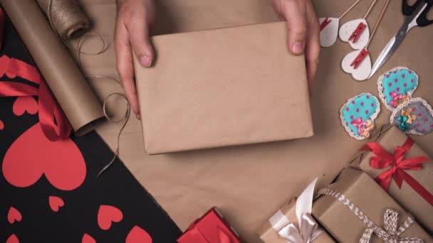 男性の手はテーブルの上にクラフト紙に箱を入れて紐で結び始めます。休日のための贈り物を準備するプロセス。バレンタインのお祝いのコンセプト - 映像、動画