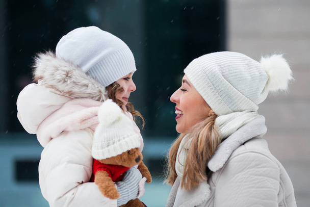 Little daughter looking at mother and holding teddy bear in winter city - Foto, Imagem