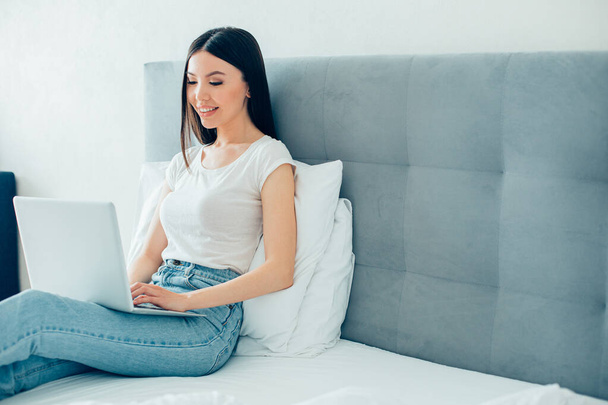 Giovane donna dai capelli lunghi seduta sul letto con un computer portatile moderno e guardando lo schermo. banner del sito web - Foto, immagini