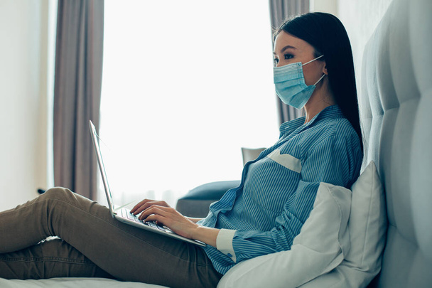 Young lady wearing a medical mask sitting on the bed with a modern laptop on her laps - Foto, Imagem