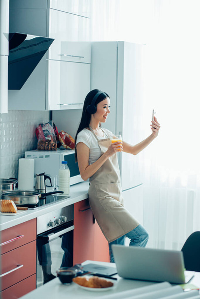 Beautiful lady in casual clothes and an apron standing with a glass of orange juice and smiling while taking photos - 写真・画像
