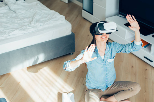 Smiling young woman sitting on the floor with hands up while using virtual reality glasses. Website banner - Foto, Imagen