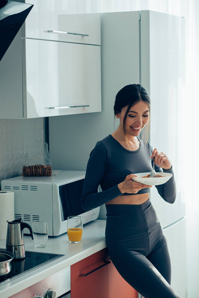 Mirthful pretty lady smiling while standing with a bowl of cornflakes in the kitchen - Foto, Imagem