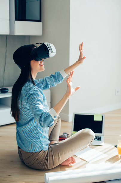 Mirthful lady sitting cross legged on the floor in front of a laptop and wearing virtual reality device - Фото, зображення