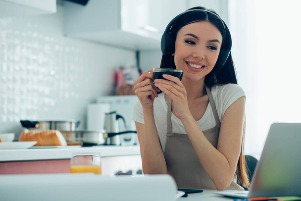 Cheerful beautiful young woman wearing headphones looking at the laptop screen with a smile while holding a cup - Photo, image