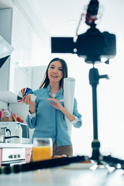 Pretty lady standing with a paper roll and color swatches while talking on the camera in her kitchen - Фото, зображення
