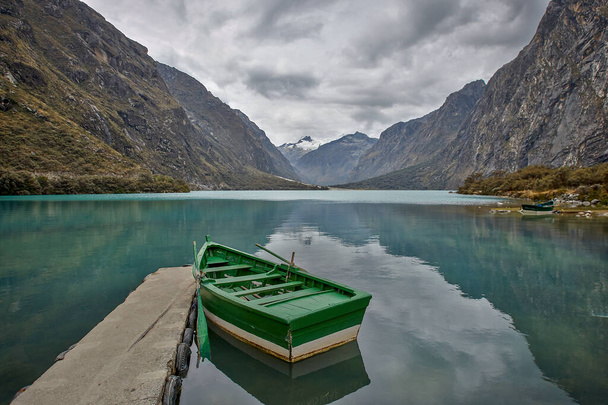 Kleines grünes Boot, das an einem kleinen Steg an einem Süßwassersee festgemacht hat, umgeben von hohen felsigen Bergen mit Schnee und wolkenverhangenem Himmel mit drohendem Regen - Foto, Bild