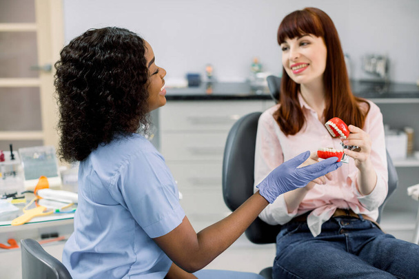 Dentist and patient in modern clinic. Smiling happy african female dentist and Caucasian girl patient holding together teeth model, speaking and looking each other. Oral care, orthodontics - Foto, immagini