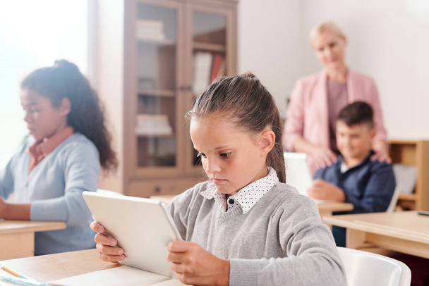 Σοβαρό χαριτωμένο κορίτσι και τους συμμαθητές της με ψηφιακές ταμπλέτες κάθεται από γραφεία και εργάζονται ατομικά με online κείμενο, ενώ ο δάσκαλος συμβουλεύεται τους - Φωτογραφία, εικόνα