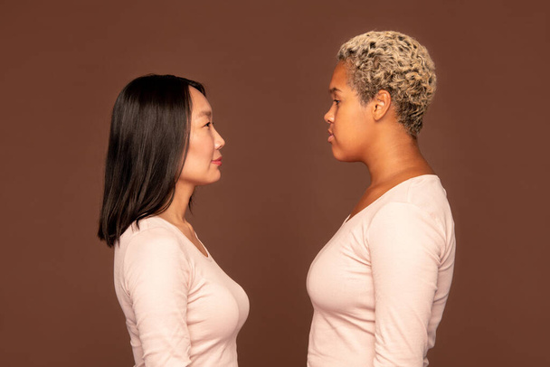 Sivunäkymä kahdesta nuoresta kulttuurienvälisestä naisesta valkoisissa pujopaidoissa, jotka seisovat kameran edessä ruskeaa taustaa vasten ja katsovat toisiaan - Valokuva, kuva
