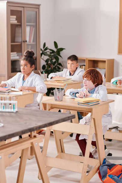 Group of elementary schoolkids in whitecoats sitting by desks in two rows at lesson of chemistry and listening to their teacher explanations - Φωτογραφία, εικόνα