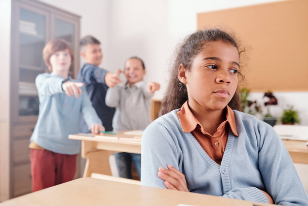 Urażona uczennica rasy mieszanej, siedząca przy biurku i przechodząca ramię w ramię na tle kolegów z klasy znęcających się nad nią podczas przerwy w klasie. - Zdjęcie, obraz