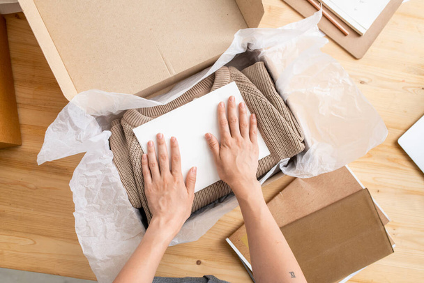 Hands of young female manager of online shop putting paper on top of folded sweater into box before packing it and sending to client - Photo, Image
