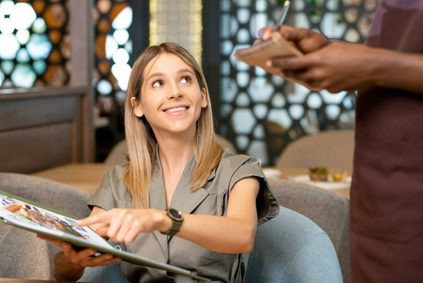 Mains de serveur africain debout à table et prenant ordre de fille souriante blonde pointant vers le menu tout en commandant un dessert ou une salade dans un café - Photo, image
