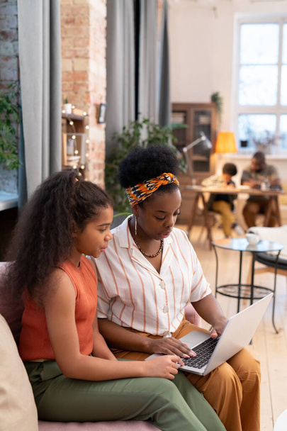 Junge Frau afrikanischer Herkunft und ihre Teenager-Tochter sitzen auf dem Sofa im Wohnzimmer und schauen Online-Unterricht gegen Vater und Sohn - Foto, Bild