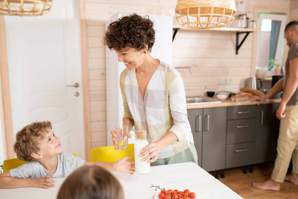 Madre feliz mirando a uno de los dos niños y poniendo una botella de leche fresca en la mesa mientras su marido cocinando el desayuno en el fondo - Foto, imagen