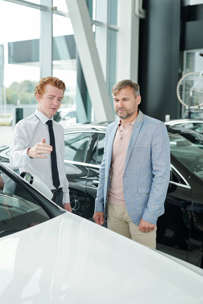 Зрелый мужчина в повседневной одежде, слушая молодого менеджера по продажам, указывая на характеристики нового белого автомобиля при выборе нового автомобиля - Фото, изображение