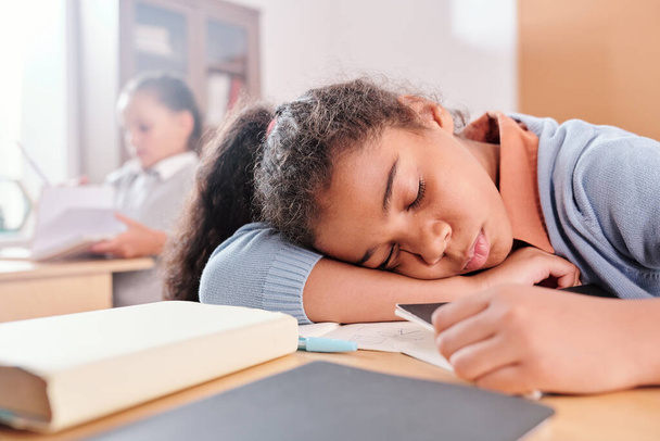 Uitgeput of verveeld schoolmeisje in casualwear houden haar hoofd op het bureau tijdens het slapen op de achtergrond van klasgenoot met boek - Foto, afbeelding