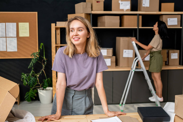 Jovem sorridente mulher trabalhador de loja on-line escritório em pé por local de trabalho contra seu colega asiático vai levar uma das caixas com ordens - Foto, Imagem