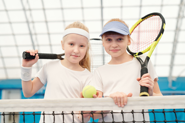 Due ragazze simpatiche amichevoli in abbigliamento sportivo bianco in piedi dalla rete allo stadio di fronte alla telecamera e tenendo racchette da tennis sulle spalle pronte per il gioco - Foto, immagini