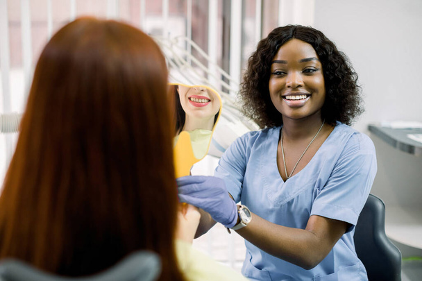 Back view of woman patient looking at her smile after curing or whitening teeth in dental clinic, and happy African female dentist, showing the mirror with reflection of smile - Foto, afbeelding