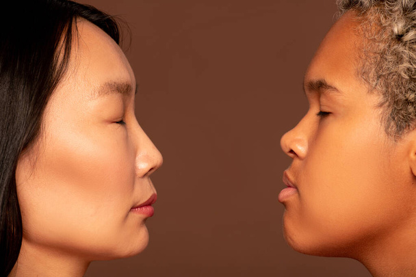 Side view of faces of two young intercultural women standing in front of camera against brown background and keeping their eyes closed - Photo, Image